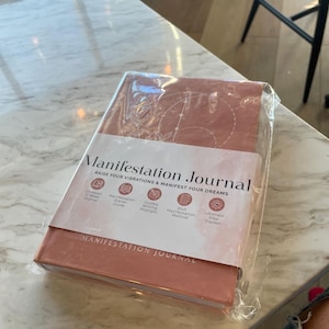 Pu&ograve; includere: Un diario di manifestazione rettangolare color terracotta avvolto in plastica trasparente. La copertina presenta disegni a linee bianche di simboli celesti e il testo "Manifestation Journal" con una fascia di carta che elenca varie caratteristiche.