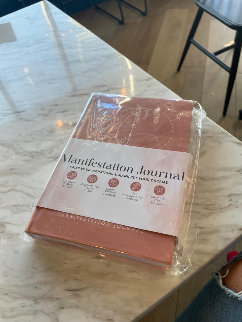 Pu&ograve; includere: Un diario di manifestazione rettangolare color terracotta avvolto in plastica trasparente. La copertina presenta disegni a linee bianche di simboli celesti e il testo "Manifestation Journal" con una fascia di carta che elenca varie caratteristiche.