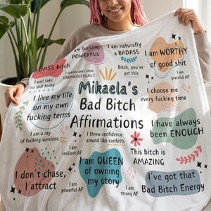 May include: A white fleece blanket featuring the text Mikaela's Bad Bitch Affirmations surrounded by colorful abstract shapes and various motivational phrases including I am naturally a BADASS, I am WORTHY of good shit, and I am resilient AF.