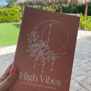 Pu&ograve; includere: Un diario color rosa antico con una grafica a linee bianche che raffigura una luna crescente, fiori e forme geometriche. La copertina riporta il testo High Vibes MANIFESTATION JOURNAL in una combinazione di caratteri bianchi e puliti.