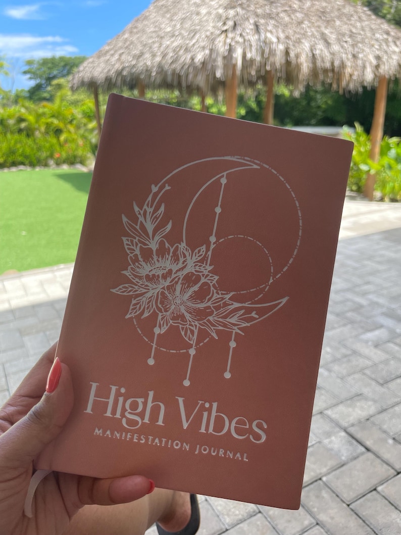 Pu&ograve; includere: Un diario color rosa antico con una grafica a linee bianche che raffigura una luna crescente, fiori e forme geometriche. La copertina riporta il testo High Vibes MANIFESTATION JOURNAL in una combinazione di caratteri bianchi e puliti.