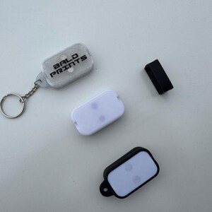 May include: Three keychains with clear plastic cases and white buttons. The keychain on the left has the text "BOLD PRINTS" on the case. The keychain on the right has a black case.