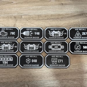 May include: A collection of black and white "Horror Badge" decals. Each badge features a unique graphic and alphanumeric code, such as "78C" and "Y46". The decals are rectangular with rounded corners and a white border.