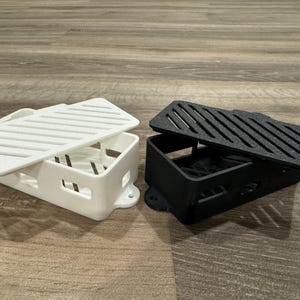 May include: Two 3D printed cases with lids, one white and one black. The cases have a grid pattern on the lids and are designed to hold small electronics.