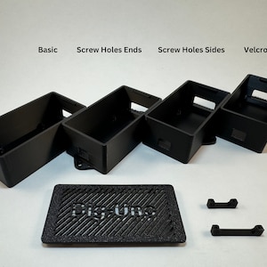 May include: Five black plastic enclosures with different mounting options, including screw holes on the ends and sides, and a velcro option. The enclosures are arranged in a row from left to right. A black plastic plate with the word "Digium" printed on it is in the bottom center of the image. Two black plastic brackets are in the bottom right corner of the image.