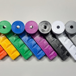 May include: A collection of colorful, 3D-printed handles in a rainbow of hues, including red, orange, yellow, green, blue, purple, gray, and white. Each handle has a circular grip and a textured, angled body.