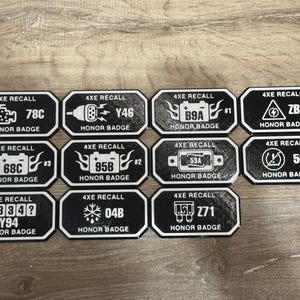 May include: A collection of black and white "4XE RECALL HONOR BADGE" stickers. Each badge features a unique code and graphic, such as a battery, engine, or snowflake. The stickers are arranged on a wooden surface.