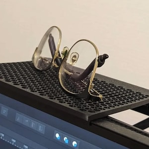 May include: A black plastic desk organizer with a honeycomb pattern. A pair of eyeglasses with gold frames and clear lenses rests on the organizer.