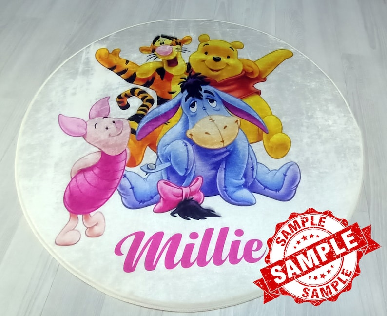 May include: A round white rug with a Winnie the Pooh theme. The rug features Winnie the Pooh, Tigger, Piglet, and Eeyore. The rug has the name "Millie" written in pink cursive letters. The rug has a red "SAMPLE" stamp on it.