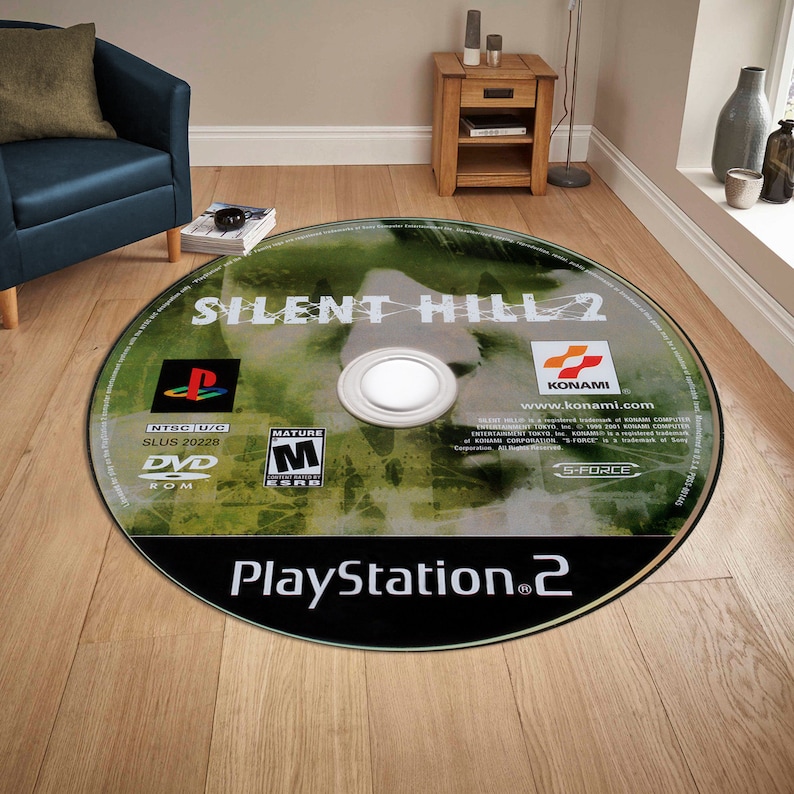 May include: A round rug featuring a close-up image of a PlayStation 2 game disc for Silent Hill 2. The disc is green and black with the game title, "Silent Hill 2", in white text. The game's rating, "Mature", is visible in the lower left corner. The game's publisher, "Konami", is visible in the upper right corner. The text "PlayStation 2" is visible in the lower center of the disc.