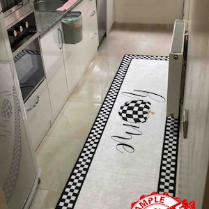 May include: A white kitchen rug with a black and white checkered border. The rug has the word "Home" written on it in black cursive with a black and white checkered pumpkin design. The rug has a red "SAMPLE" stamp on the bottom right corner.