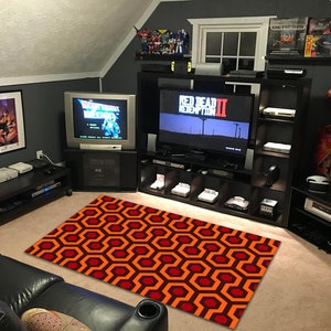 Könnte beinhalten: Ein Spielzimmer mit einem schwarz-orangen Teppich, einem großen Flachbildfernseher und einem schwarzen Unterhaltungszentrum mit Regalen voller Videospiele und Konsolen. Der Fernseher zeigt das Spiel "Red Dead Redemption II".