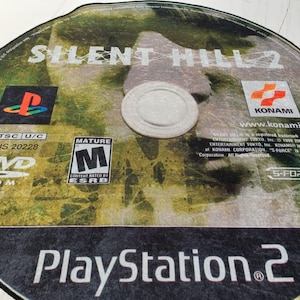 May include: A PlayStation 2 game disc for Silent Hill 2. The disc is silver with a black border and features a green and brown background with the game title in white text. The disc also includes the ESRB rating of Mature, the Konami logo, and the text "www.konami.com".