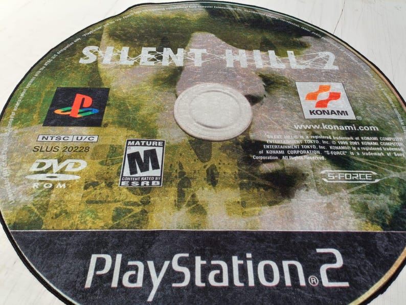 May include: A PlayStation 2 game disc for Silent Hill 2. The disc is silver with a black border and features a green and brown background with the game title in white text. The disc also includes the ESRB rating of Mature, the Konami logo, and the text "www.konami.com".