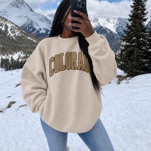 May include: A beige crewneck sweatshirt featuring the word COLORADO printed in a large, arched, gold-colored font with a dark outline across the chest. The garment has long sleeves with ribbed cuffs and a ribbed neckline, providing a relaxed fit.