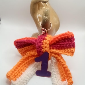 May include: A handmade crochet bow keychain in shades of orange, red, and white, with a purple number "1" charm. The keychain has a copper-colored ring. The bow is a decorative accessory.
