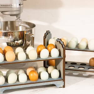May include: Two wooden egg holders with blue paint and brown trim. The holders are filled with brown and white eggs. The holders are on a white countertop next to a silver stand mixer.