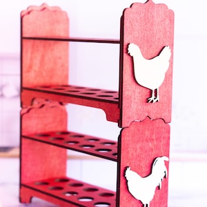 May include: A red wooden egg holder with two shelves and a rooster and hen design. The holder has 12 slots for eggs.