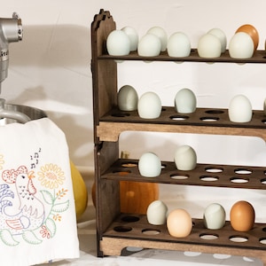 May include: A wooden egg storage rack with four shelves, each holding rows of white and brown eggs. The rack is made of dark wood and has a rustic look. The rack is sitting on a white countertop next to a silver stand mixer.