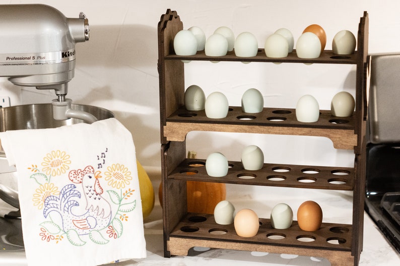 May include: A wooden egg storage rack with four shelves, each holding rows of white and brown eggs. The rack is made of dark wood and has a rustic look. The rack is sitting on a white countertop next to a silver stand mixer.
