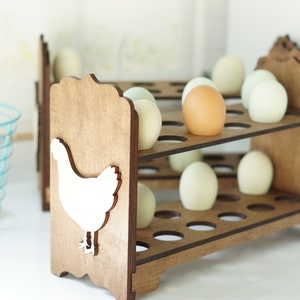 May include: A wooden egg holder with a chicken silhouette on the side. The holder has two tiers and can hold 18 eggs. The eggs are a mix of white and brown.