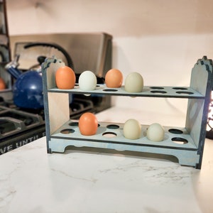 May include: A two-tiered wooden egg holder with a light blue finish. The holder has 18 individual slots for eggs. There are six eggs in the holder, three white and three brown.