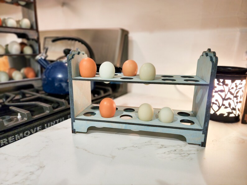 May include: A two-tiered wooden egg holder with a light blue finish. The holder has 18 individual slots for eggs. There are six eggs in the holder, three white and three brown.