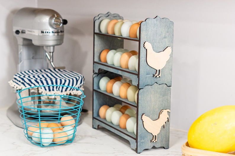 May include: A three-tiered wooden egg holder with a light blue finish. Each tier holds six eggs. The holder features a cutout of a chicken on each side. The holder is filled with a variety of brown and white eggs.