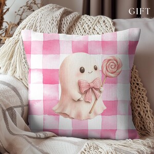 May include: A square decorative pillow with a pink and white gingham pattern. The pillow features a watercolor illustration of a friendly ghost holding a lollipop and wearing a bow. The text "GIFT IDEA" is in the upper right corner.