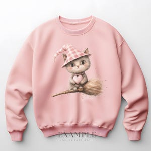 May include: A light pink sweatshirt featuring a whimsical illustration of a gray kitten wearing a pink and white checkered witch hat and holding a pink heart. The kitten is sitting on a broom. The text "EXAMPLE" and "THE_CLIPART_WAY" are at the bottom.