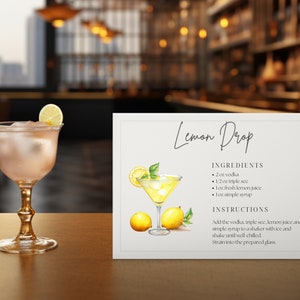 May include: A refreshing Lemon Drop cocktail in a stemmed glass, garnished with a lemon slice, next to a sign with the recipe. The sign includes the ingredients: vodka, triple sec, lemon juice, and simple syrup. A small candle holder is also visible.