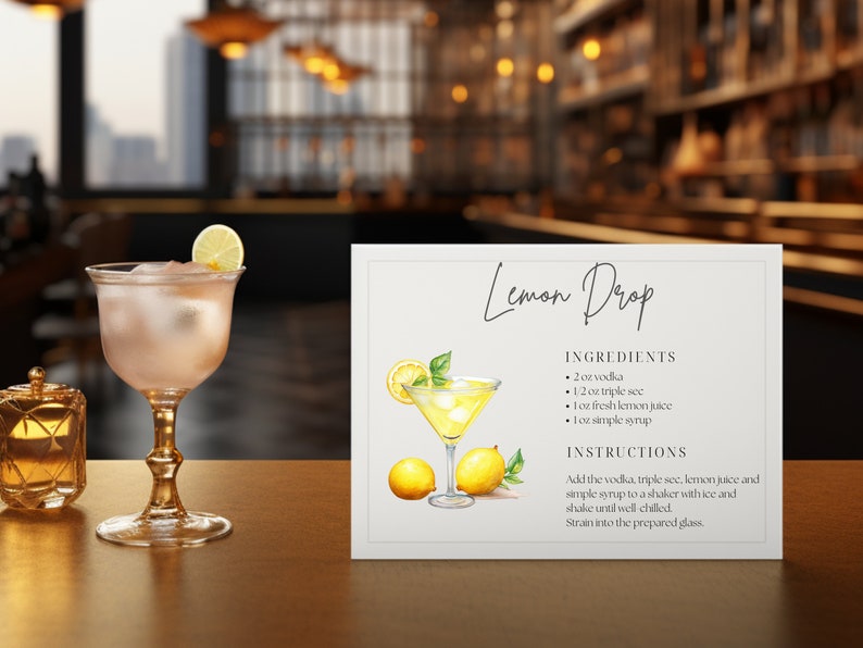 May include: A refreshing Lemon Drop cocktail in a stemmed glass, garnished with a lemon slice, next to a sign with the recipe. The sign includes the ingredients: vodka, triple sec, lemon juice, and simple syrup. A small candle holder is also visible.