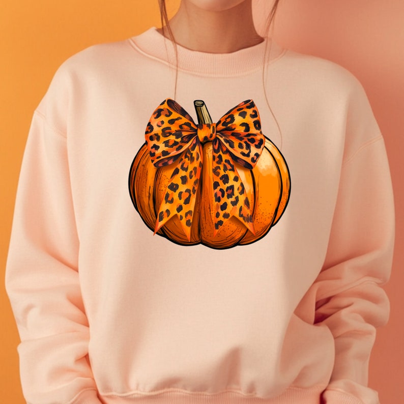 May include: A light pink sweatshirt with a graphic of an orange pumpkin with a leopard print bow.