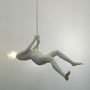 May include: A white, textured, human-shaped sculpture hangs from the ceiling with a light bulb in its head. The sculpture is suspended by a thin cord and is in a relaxed, hanging position.