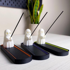 Puede incluir: Tres porta inciensos blancos con forma de fantasma con varillas de incienso negras. Cada fantasma se asienta sobre una bandeja rectangular negra con ribete de colores: naranja, morado y verde. Los soportes están sobre una superficie blanca.