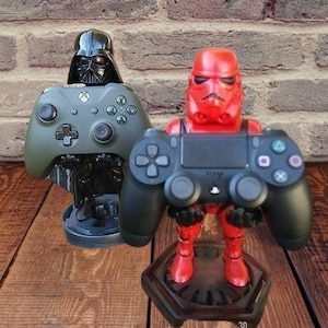 May include: Two video game controller stands, one shaped like Darth Vader and the other shaped like a Stormtrooper. Both stands are black and have a red base.