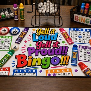 May include: A rectangular bingo game mat featuring the text Yell it Loud Yell it Proud!! Bingo!!! in colorful lettering, surrounded by bingo cards, numbered balls, and several colorful bingo daubers. The set includes a wire cage for balls.
