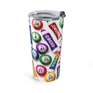 May include: A white travel tumbler featuring a colorful bingo-themed design. The tumbler is covered with images of bingo balls and bingo markers in various colors, including red, green, purple, and yellow. The tumbler has a clear lid and a black slider.