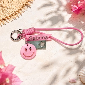 Könnte beinhalten: Ein Schlüsselanhänger aus rosa Paracord mit einem silbernen Metallring, einem rosa rechteckigen Anhänger mit dem in Schwarz gedruckten Namen Sabrina, einem dunkelgrünen rechteckigen Anhänger mit Sternausschnitt und einem runden rosa Smiley-Anhänger.