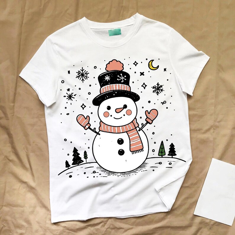 May include: White t-shirt with a cartoon snowman illustration. The snowman has a black top hat, a pink and white striped scarf, and pink mittens. Snowflakes and a crescent moon are also shown in the design.