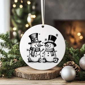 May include: A white, circular Christmas ornament with a black and white illustration of two snowmen wearing top hats and scarves. The design includes snowflakes and small cups. A cream ribbon is attached for hanging.