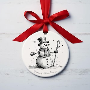May include: A white Christmas bauble featuring a black and white drawing of a snowman in a top hat, scarf, and gloves, holding a cane. It's tied with a red ribbon and has the text "Merry Christmas!"