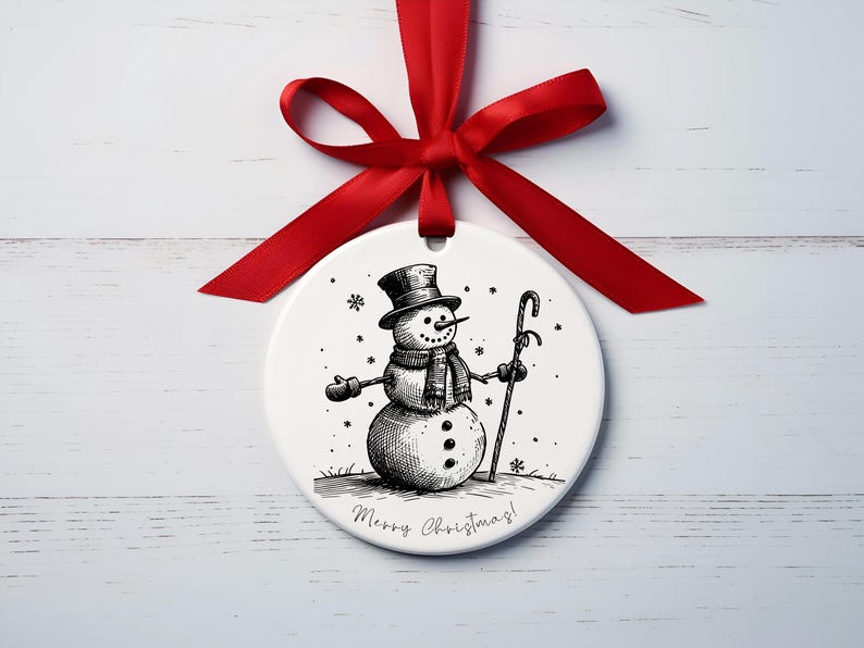 May include: A white Christmas bauble featuring a black and white drawing of a snowman in a top hat, scarf, and gloves, holding a cane. It's tied with a red ribbon and has the text "Merry Christmas!"