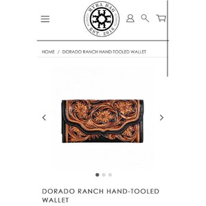 Puede incluir: Una cartera de cuero negra y marrón con estampado floral. La cartera está cerrada y tiene una solapa con un detalle de costura decorativa. La cartera está sobre un fondo blanco.