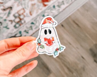 May include: A white ghost wearing a red Santa hat and holding a red book. The ghost is surrounded by green holly leaves and red berries.