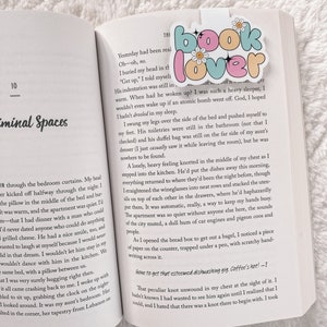 Może przedstawiać: Zbliżenie otwartego książki z r&oacute;żowo-niebieskim zakładką z napisem "book lover" z kwiatami i błyskotkami. Tekst na stronie brzmi "Yesterday had been real. Oh-oh, no. I buried my head in the..."