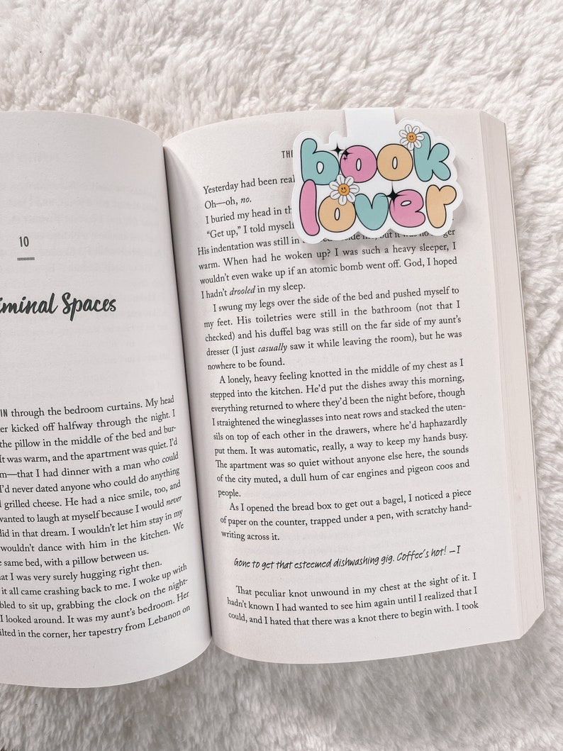 Może przedstawiać: Zbliżenie otwartego książki z r&oacute;żowo-niebieskim zakładką z napisem "book lover" z kwiatami i błyskotkami. Tekst na stronie brzmi "Yesterday had been real. Oh-oh, no. I buried my head in the..."