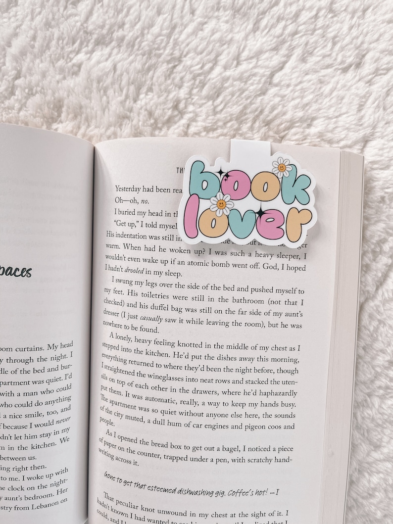 Może przedstawiać: Zbliżenie na zakładkę z napisem "Book Lover" w kolorze r&oacute;żowym, niebieskim i zielonym. Zakładka zawiera napis "Book Lover" z motywem stokrotki i błyszczącymi akcentami. Zakładka znajduje się na otwartej książce z tekstem o porannej rutynie osoby.