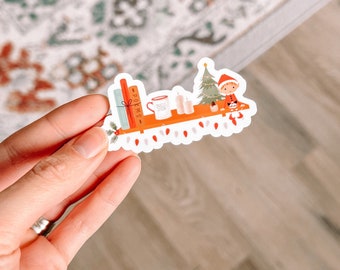May include: A sticker featuring a small elf sitting on a shelf with a Christmas tree, a mug, candles, and books. The shelf is decorated with white lights. The sticker is on a white background.
