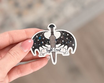 Op de afbeelding: Een zwart-witte sticker met een zilveren zwaard en vleugels met sterren. De sticker heeft de tekst "Night Court" op een banner.